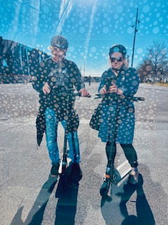Mann und Frau auf E-Roller mit Helmen auf dem Kopf, fotografiert in einer spiegelnden Scheibe vor tiefblauem Himmel