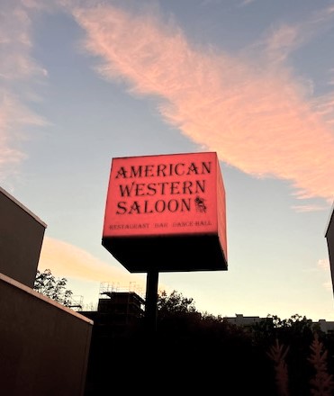 Leuchtschild des American Western Saloon mit den Worten 'AMERICAN WESTERN SALOON', 'RESTAURANT BAR DANCE HALL' in schwarzer Schrift auf einem quadratischen, roten, beleuchteten Schild vor Himmel bei Sonnenuntergang.