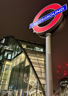 Rundes Londoner Underground-Schild mit rotem Rand und blauer Schrift 'UNDERGROUND', vor einem modernen Gebäude bei Nacht.
