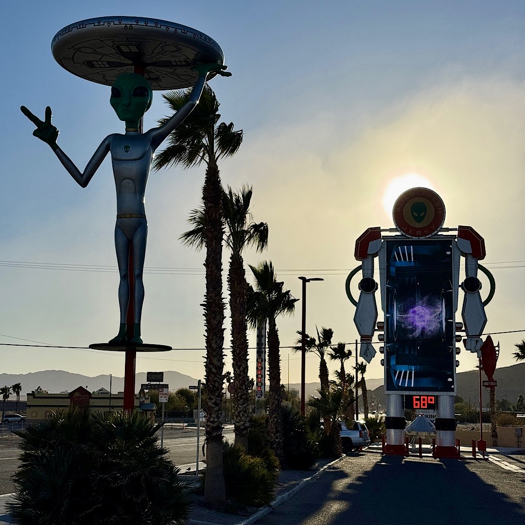 Silouette einer überlebensgroßen silberner Alienfigur mit einem runden UFO auf dem Kopf, daneben eine große Roboterfigur mit einem Bildschirm, der 68 Grad anzeigt, daneben hohe Palmen im Gegenlicht. Hinter dem Roboter geht die Sonne unter, der Himmel ist klar und hellblau