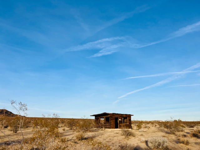 Holzhütte in einer weiten, wüstenartigen Landschaft mit einigen trockenen Büschen unter blauem, klarem Himmel
