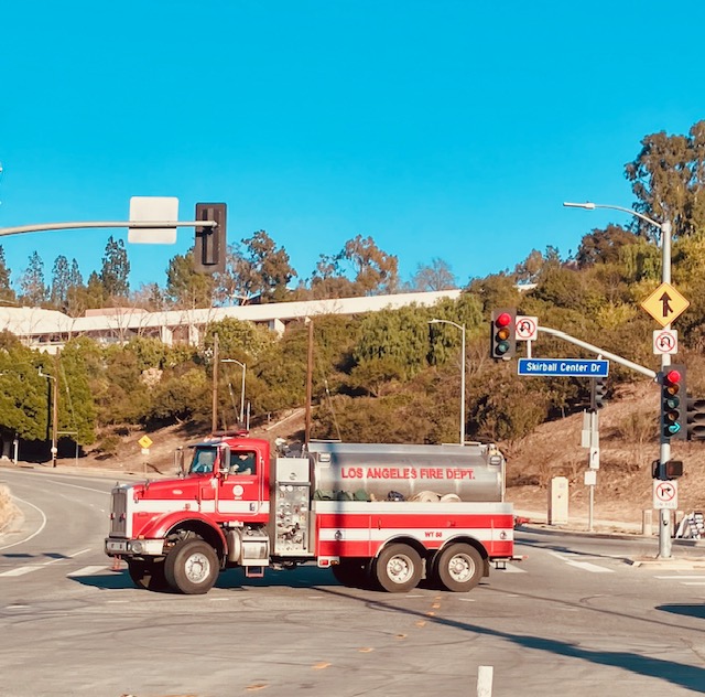 Rotes Feuerwehrfahrzeug des Los Angeles Fire Department an einer Kreuzung. Verkehrssignale und Straßenbeschilderung sichtbar. Im Hintergrund eine Böschung mit Sträuchern unter klarem, blauem Himmel