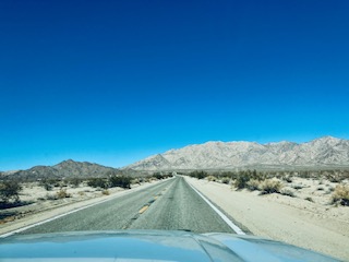 Blick aus dem Auto auf eine gerade Straße, die durch eine Wüstenlandschaft führt, mit Bergen im Hintergrund und klarem blauen Himmel.