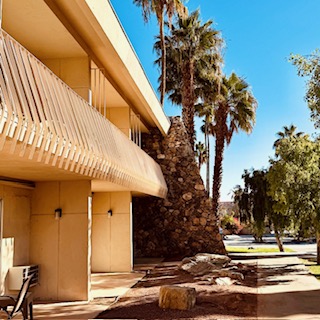 Zweistöckiges Motelgebäude im Retro-Stil mit Balkonen, hellbeiger Fassade, weißen Geländern, Palmen im Hintergrund unter strahlend blauem Himmel