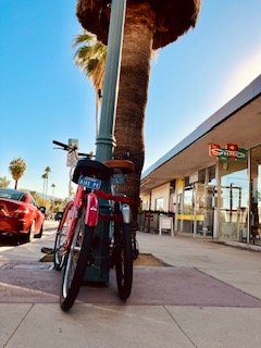 Zwei Fahrräder stehen an einem Laternenpfahl vor einem Geschäft in Palm Springs. Im Hintergrund sind Palmen und ein Auto sichtbar. Der Himmel ist klar und hellblau, die Fährrader sind von hinten zu sehen
