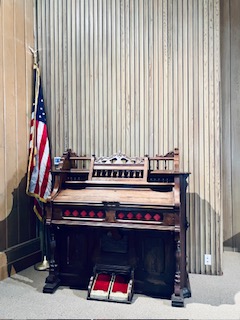 Schlichte, altmodische Kirchenorgel aus Holz mit roten und schwarzen Tasten, neben einer zusammengerollten US-Flagge, vor einer Wand mit vertikaler Holzverkleidung.