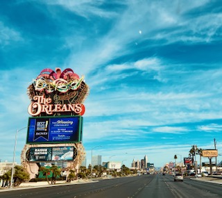Üppig dekoriertes Retro-Schild des Hotels 'The Orleans' an einer Straße in Las Vegas. Im Hintergrund ist die Skyline des Las Vegas Strips zu sehen unter blauem Himmel mit freundlichen weißen Wolken