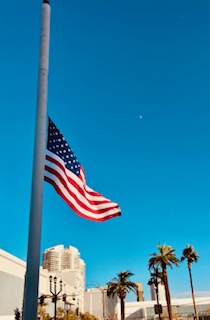 Die US-Flagge weht auf Halbmast an einer Fahnenstange, im Hintergrund Palmen und eine Stadtlandschaft unter klarem, tiefblauem Himmel