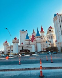 Blick auf die einem Märchenschloss nachempfundene Hotelanlage "Magic Kingdom" in Las Vegas mit vielen weißen Türmen mit spitzen roten und weißen Dächern vor klarem blauem Himmel, im Vordergrund orange-weiß-rot gestreifte Verkehrspoller