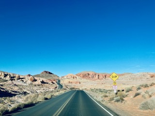 Eine gerade asphaltierte Straße führt durch die Wüste mit roten Felsen im Hintergrund. Ein gelbes Verkehrsschild mit der Aufschrift 'DIP' steht am Straßenrand unter klarem, tiefblauem Himmel