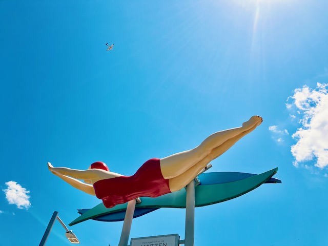 Überlebensgroße Statue einer Schwimmerin in rotem Retro-Badeanzug mit roter Badekappe, die in einer Sprungposition über zwei grüne Surfbretter ausgestreckt ist vor tiefblauem Himmel mit wenigen, sonnenbeschienenen Wolken