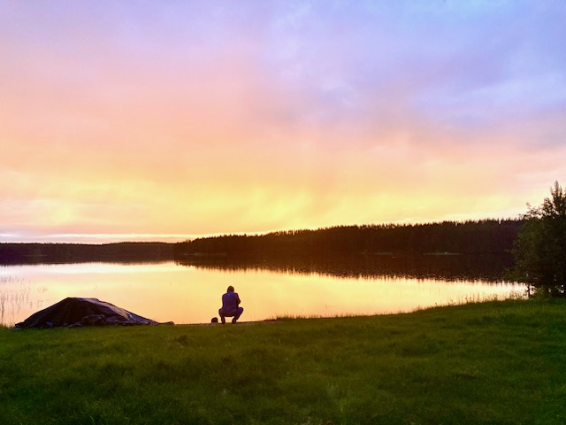 Eine Person kauert am Ufer eines ruhigen Sees bei Sonnenuntergang. Der Himmel ist in dramatische Gelb- Orange- und Lilatönen gefärbt. Aufnahme im Gegenlicht
