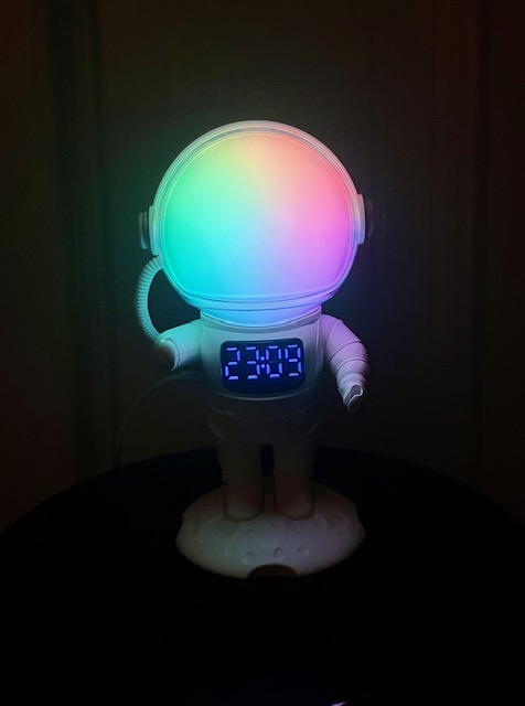 Ein Astronautenwecker mit einem bunten, leuchtenden Visier und einer digitalen Uhranzeige, die '23:09' zeigt. Der Astronaut steht auf einer kleinen Mondbasis vor dunklem Hintergrund.