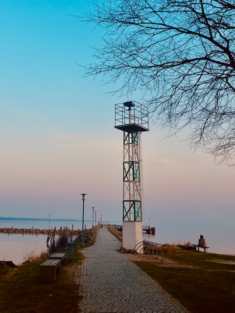 Ein viereckiger Leuchtturm mit Gitterstruktur steht am Ende eines gepflasterten Weges, der zum Wasser führt. Im Vordergrund Äste eines kahlen Baumes vor blassblauem Himmel im Sonnenuntergang