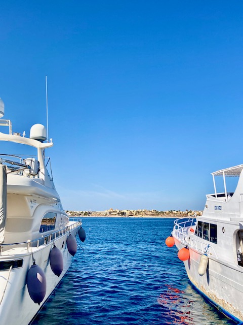 Zwei weiße Yachten stehen mit Abstand nebeneinander im Wasser, dazwischen tiefblaues Meer unter klarem, tiefblauem Himmel, im Hintergrund eine Felsenküste