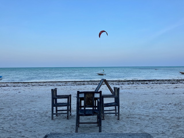 Vier Holzstühle und ein Holztisch stehen am Strand von Jambiani im Sand, Abenddämmerung, türkisfarbenes Meer, am klaren, hellblau-rosa Himmel das Segel eines Kitesurfers