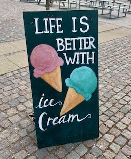 Schwarze, handbemalte Tafel mit der Aufschrift 'Life is better with ice cream' und zwei Eistüten, eine mit rosa und eine mit blauem Eis
