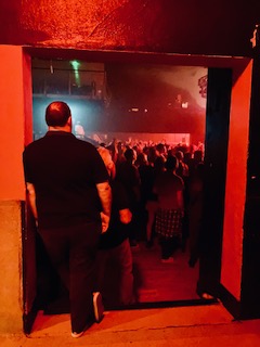 Männliche Person in dunkler Kleidung steht im Türrahmen und blickt auf eine Menschenmenge bei einem Konzert im Electric Ballroom in Camden, London, in rotes Licht getaucht