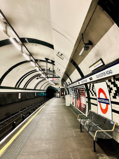 U-Bahnsteig der Station Gloucester Road in London. Sichtbare Fliesenmuster, Schienen, Bank und Schilder mit 'Gloucester Road'.