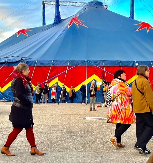 Großes blaues Zirkuszelt mit roten und gelben Stern- und Zickzackmustern, Menschen gehen davor nach rechts