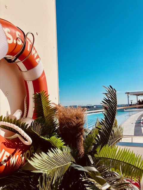 Rettungsring mit Aufschrift 'S.S. TWA New York' an Wand, davor grüne Palmblätter, blauer Himmel und Pool im Hintergrund