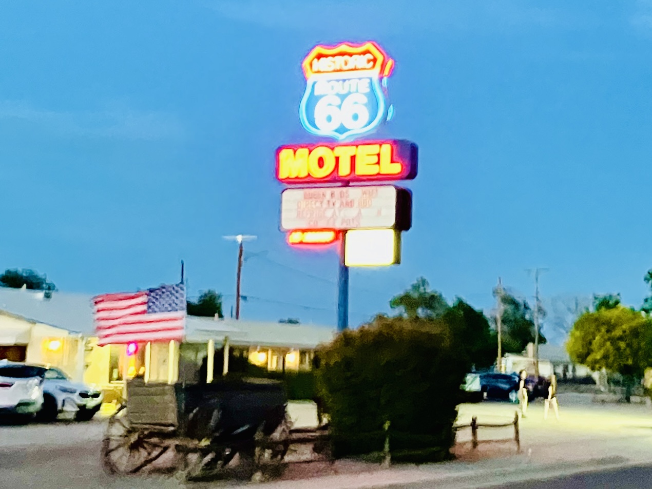 Leuchtschild mit Schrift 'Historic Route 66 Motel' und einer altmodischen Kutsche mit amerikanischen Flagge in der Abenddämmerung unter hellblauem Himmel. Dynamisch verwackelte Aufnahme