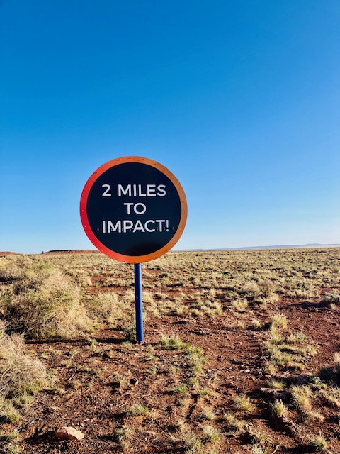 Großes, rundes Schild mit der Aufschrift '2 MILES TO IMPACT!' in einer Wüstenlandschaft unter tiefblauem Himmel