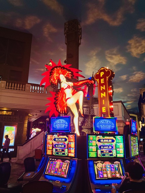 Grellbunt erleuchtende Spielautomaten mit einer großen, federbekleideten Showgirl-Figur in einem Casino, Decke ist einem künstlichen Himmel mit Wolken und Dämmerung nachempfunden