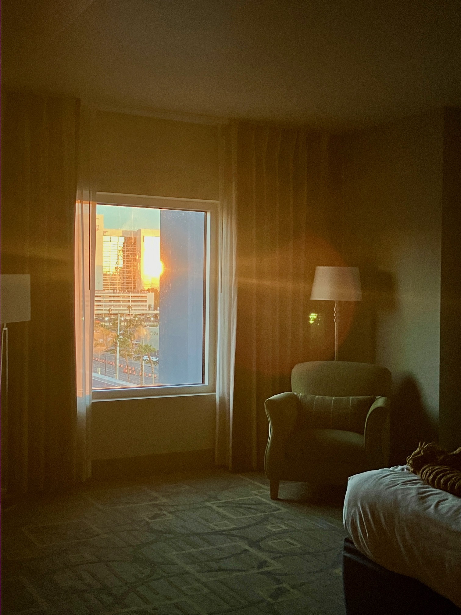 Blick aus dem Fenster eines Hotelzimmers mit einem Sessel, Stehlampe und Bett auf die verspiegelte Fassade eines Hotels in Las Vegas, die aufgehende Sonne reflektiert sich darin