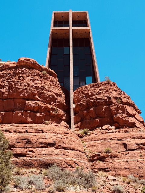 Schmale, hohe Kapelle im modernistischen Baustil aus rotem Stein mit großen Fenstern und Kreuz in der Fassade ragt zwischen roten Felsen steil in einer Wüstenlandschaft vor tiefblauem Himmel empor