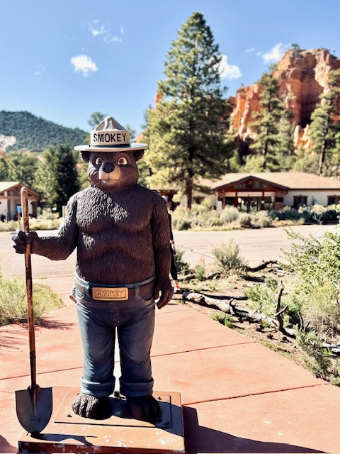 Überlebensgroße Statue von Smokey Bear mit Hut und Schaufel vor Wald und roten Felsen unter blauem Himmel