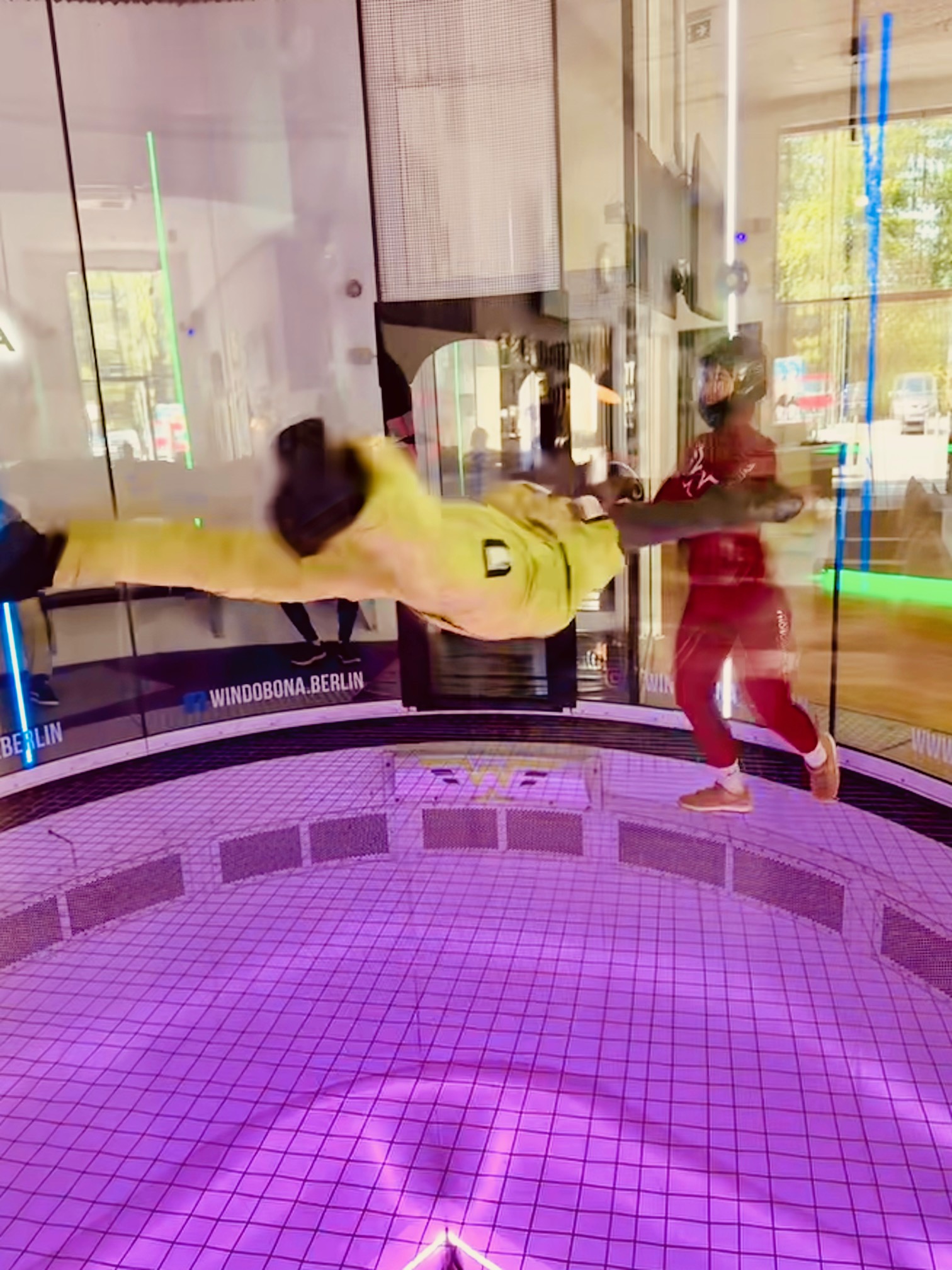 Person in gelbem Anzug schwebt horizontal im Windkanal, betreut von Trainer in rotem Anzug mit Helm