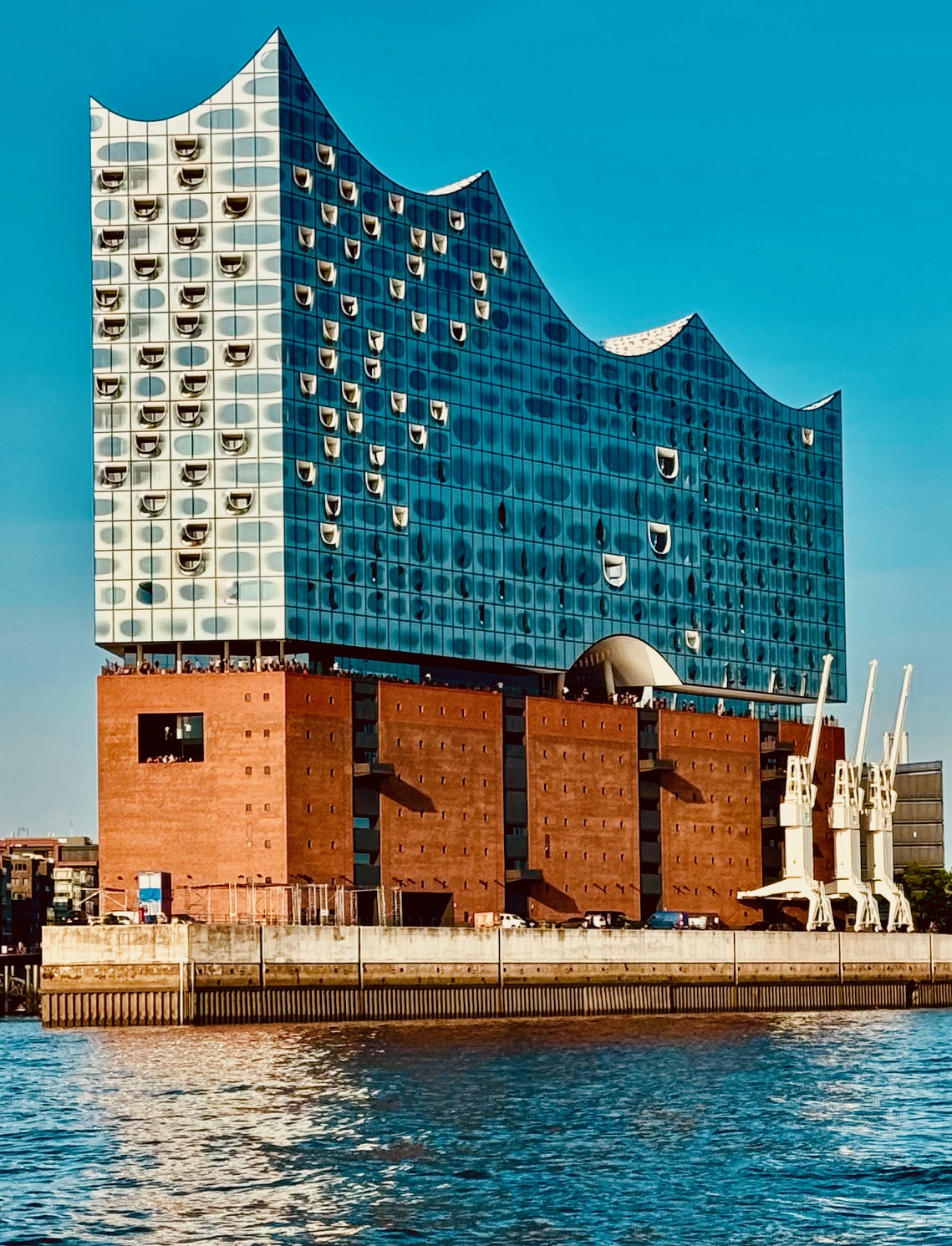 Markantes Gebäude mit rotem Backsteinsockel und silberglänzendem, wellenförmigen Aufbau, umgeben von Wasser. Außenansicht der Elbphilharmonie Hamburg