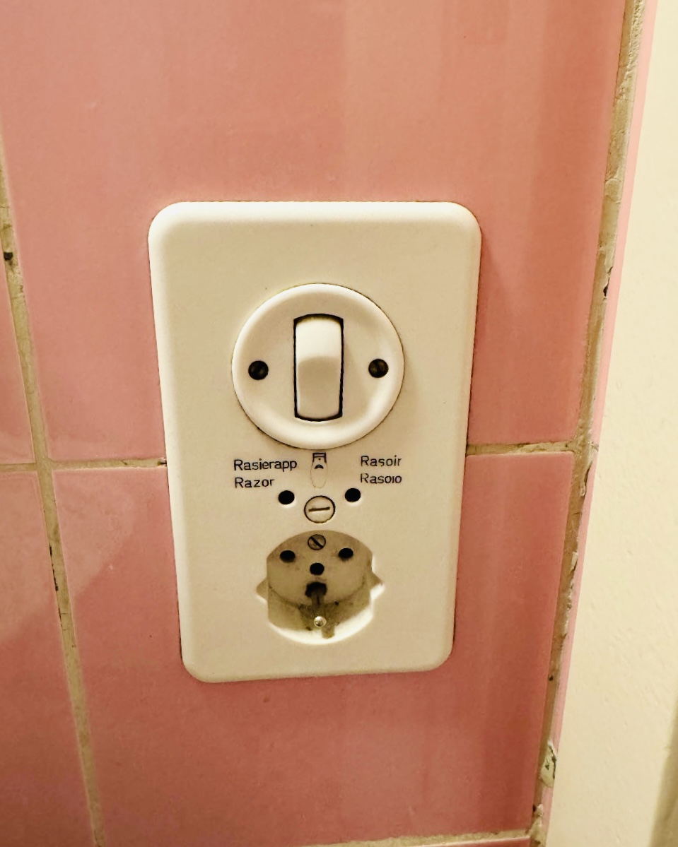 Altmodische weiße Steckdosen- und Schalterkombination an rosa gefliester Wand, Schalter für Rasierapparat mit Beschriftung in Deutsch, Englisch, Französisch und Italienisch