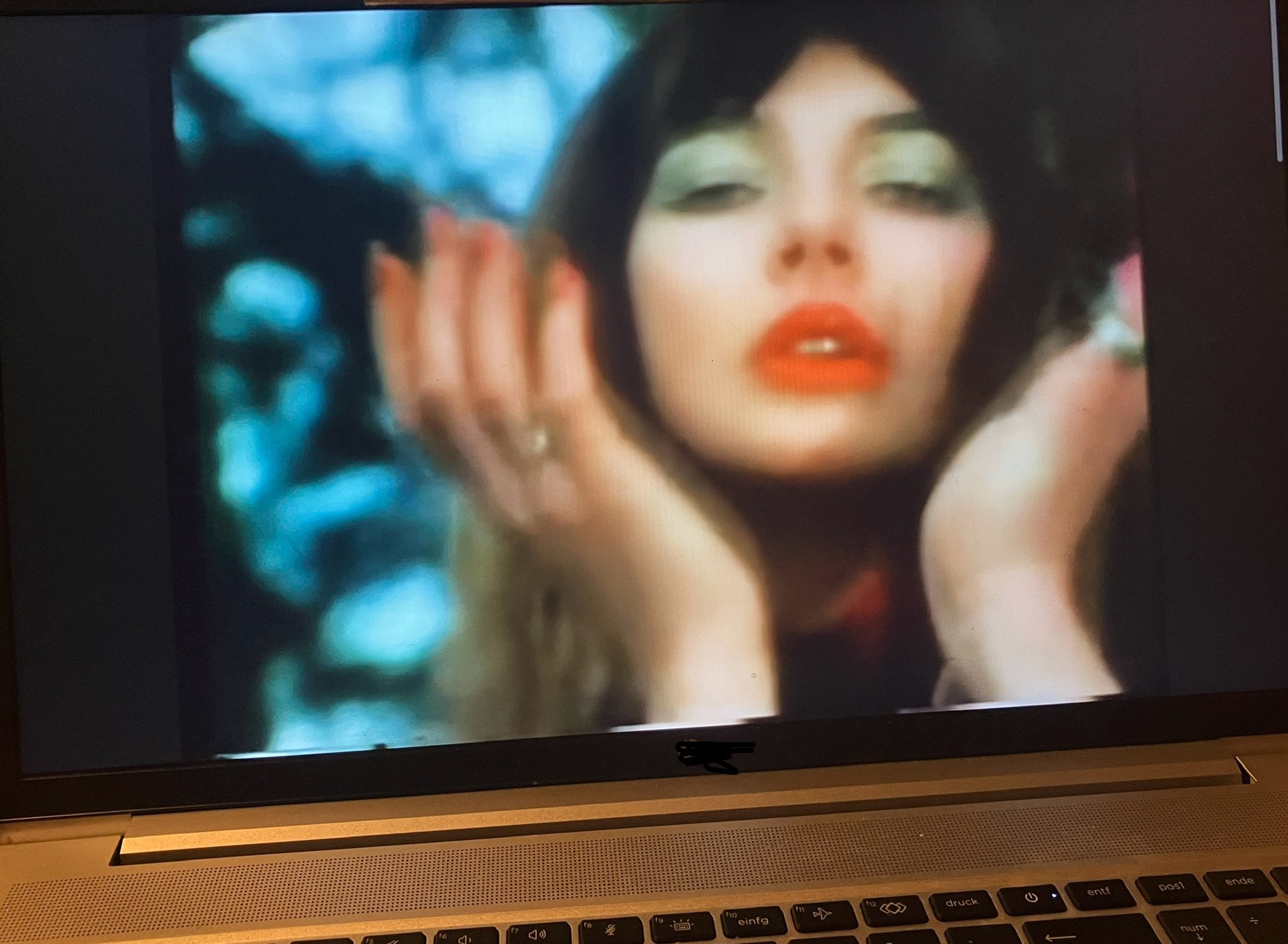 Porträt einer weiblichen Person mit dunklen Haaren, die mit den Händen von unten ihr Gesicht umrahmt vor den unscharfen Ästen eines kahlen Baums, blauer Himmel scheint durch. Die Person ist die Sängerin Kate Bush.