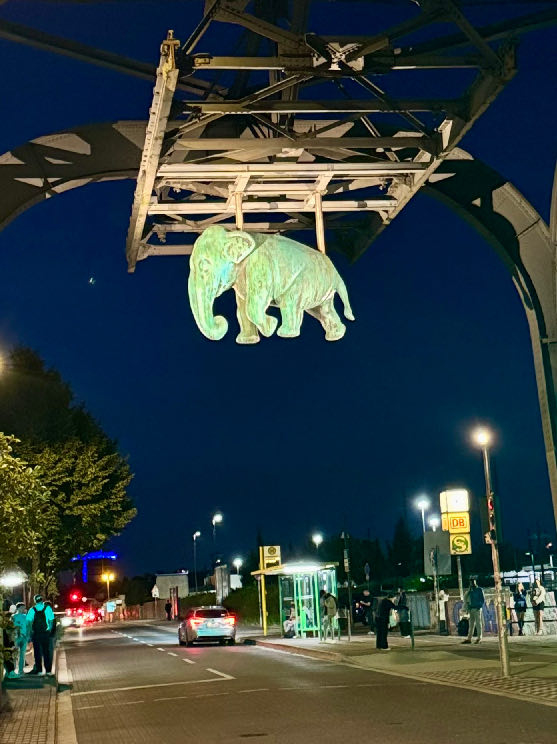 Großer hängender Elefant aus Metall mit grüner Patina an einer Straßenbahnbrücke bei Nacht in Oberhausen