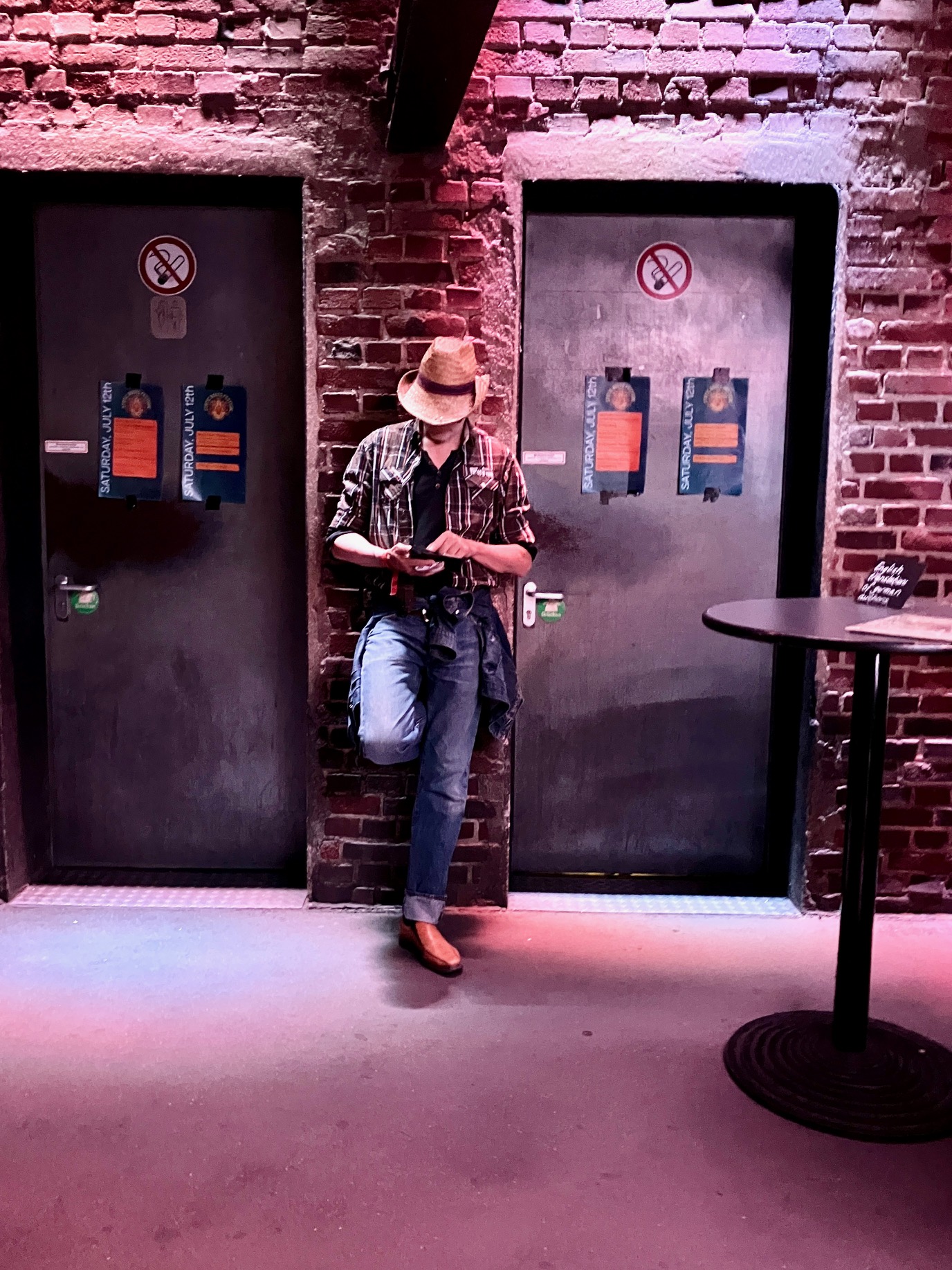 Männliche Person mit Cowboyhut, kariertem Hemd und Jeans lehnt lässig an Backsteinwand zwischen zwei Türen mit Rauchverbotsschildern. Innenreum mit schummrigem Licht.