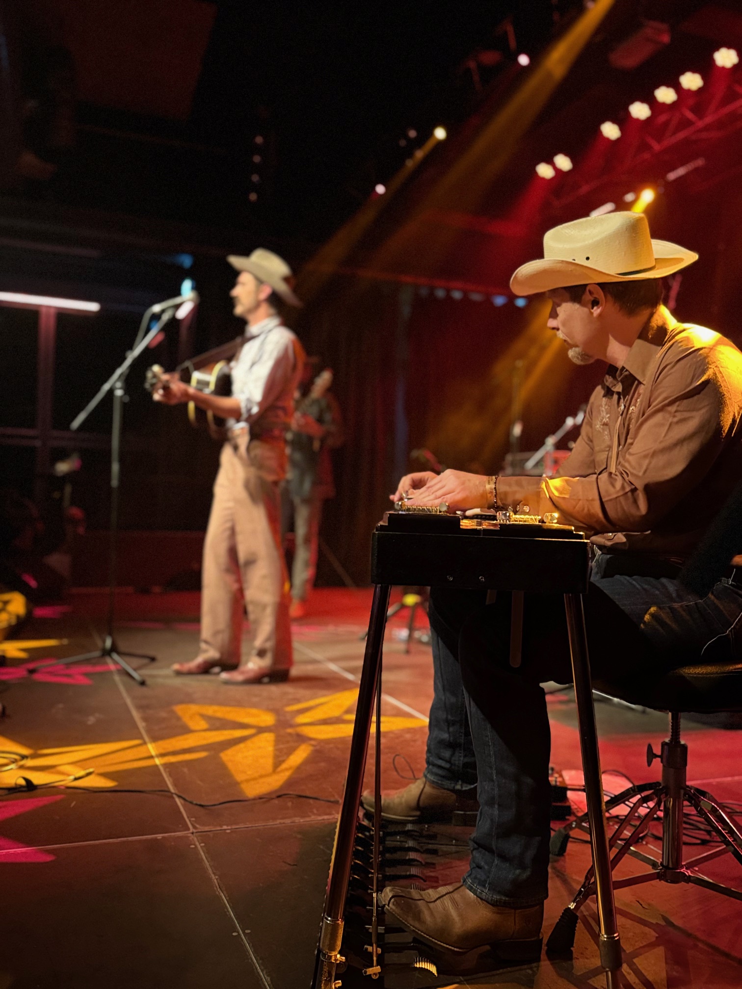 Musiker mit Cowboyhut und Westernstiefeln spielt auf einer Pedal-Steel-Gitarre auf einer Bühne sitzend mit Bühnenbeleuchtung, im Hintergrund ein stehender Mann mit Cowboyhut und Gitarre