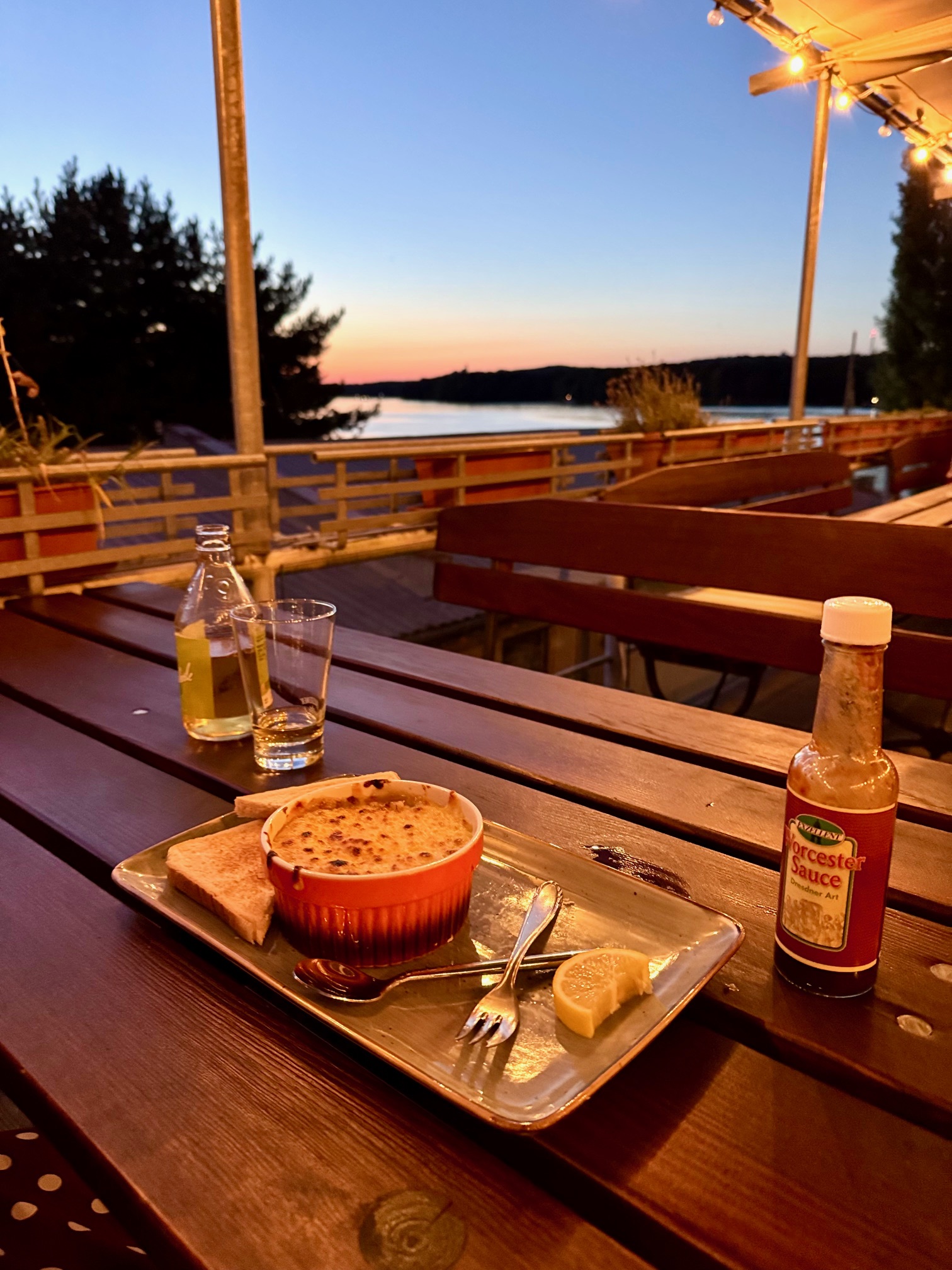 Romantische Abendstimmung an der Dahme in Berlin mit Blick auf Wasser und Sonnenuntergang, Tisch mit kleiner Auflaufform, Brot, Zitrone, Glas, Flasche und Worcestersauce