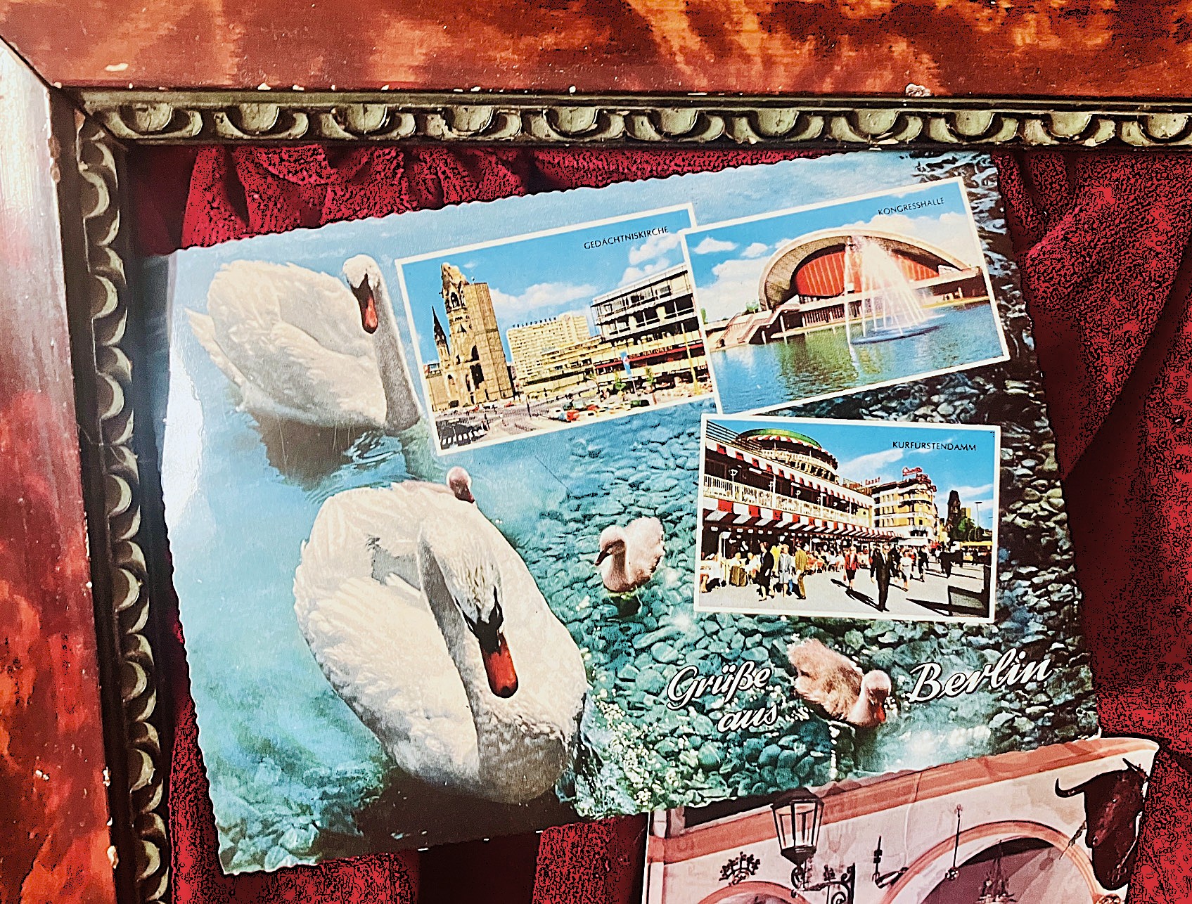 Bilderrahmen mit historischer Postkarte aus Berlin, darauf Schwäne auf Wasser, die Kaiser-Wilhelm-Gedächtniskirche, Kurfürstendamm mit Café Kranzler und Kongresshalle. Text: 'Grüße aus Berlin'.