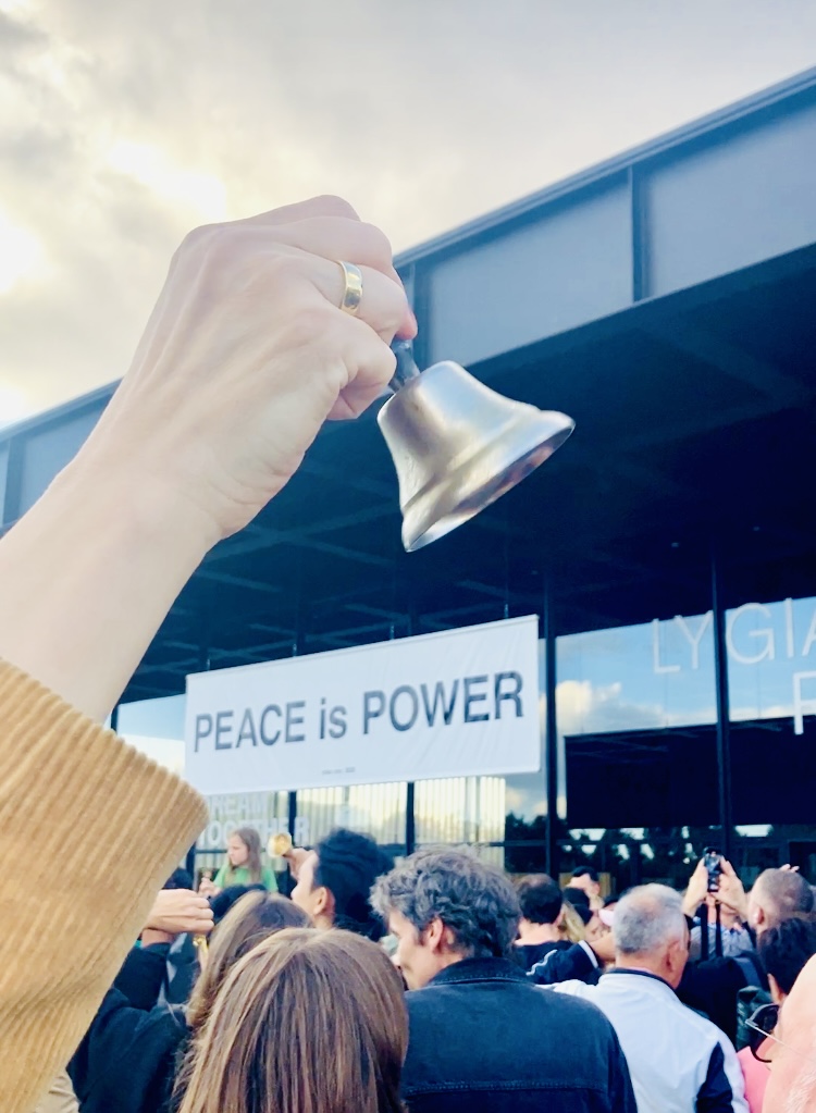 Eine Hand hält eine kleine silberne Glocke in die Höhe. Im Hintergrund ist ein Banner mit dem Text 'PEACE is POWER' zu sehen. Eine Menschenmenge versammelt sich