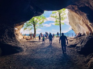 Blick aus einer Höhle auf mehrere Menschen, die draußen auf einem Weg zwischen Bäumen stehen, im Hintergrund Berge und blauer Himmel mit freundlichen Wolken
