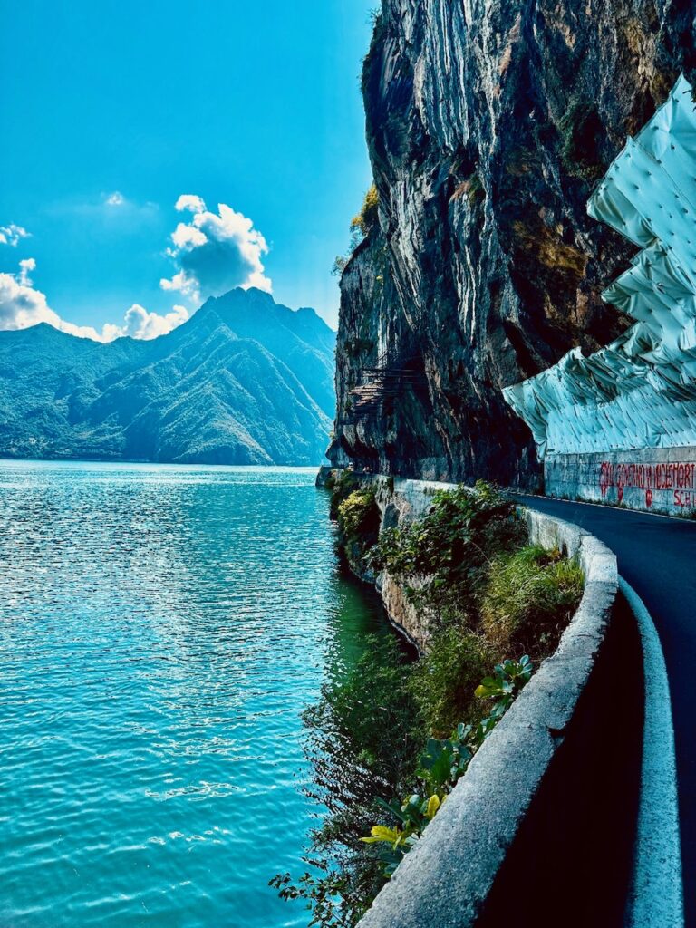 Straße am Rand eines Sees, darüber steile, überhängende Felsen, im Hintergrund bewaldete Berge unter blauem Himmel