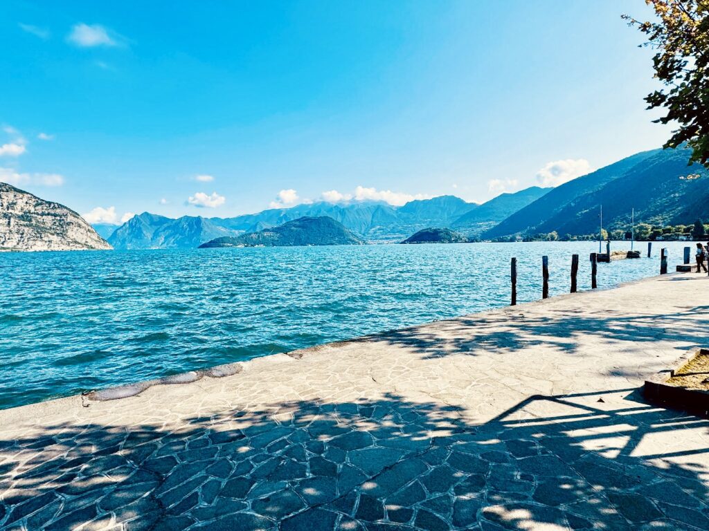 Blick auf den Lago Iseo mit blauem Wasser, bewaldeten Bergen im Hintergrund und einem steinernen Uferweg im Vordergrund unter strahlend blauem Himmel