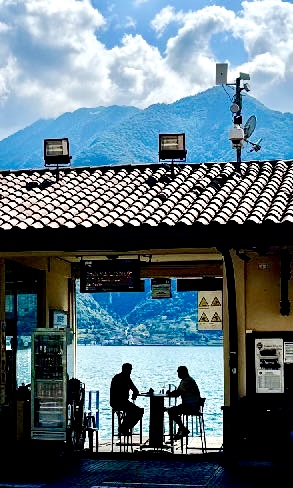 Silhouetten von zwei Personen, die an einem Tisch am Rand eines Sees sitzen, fotografiert durch eine Türöffnung, im Hintergrund bewaldete Berge