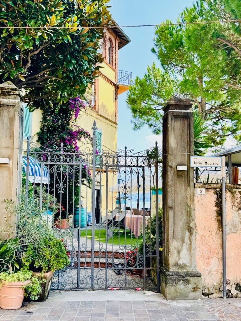 Eingang mit schmiedeeisernem Tor zu einem Garten mit Pflanzen, links ein gelbes, mediterranes Haus, davor bepflanzte Blumentöpfe, im Hintergrund der Lago Iseo