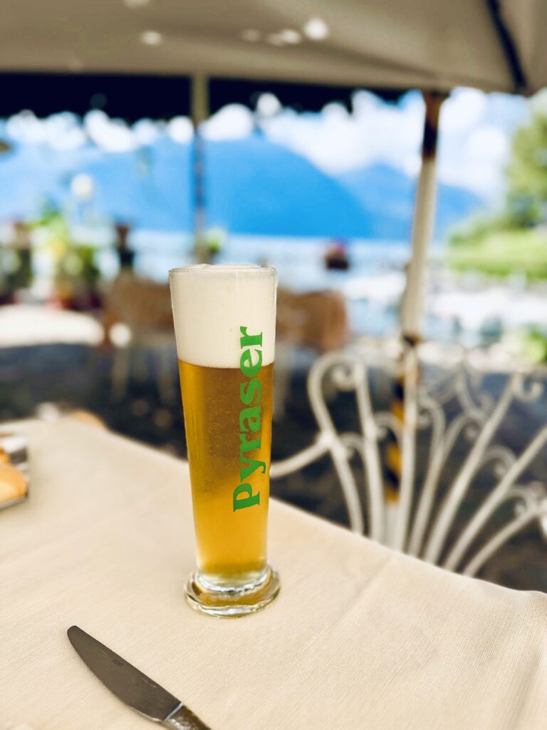 Ein Glas mit Bier steht auf einem Tisch mit weißer Tischdecke, im Hintergrund verschwommen ein See und Berge