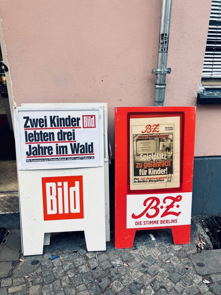 Zwei auf dem Gehweg stehende Zeitungsständer mit Schlagzeilen der Zeitungen Bild und B.Z. Die eine Schlagzeile lautet 'Zwei Kinder lebten drei Jahre im Wald', die andere Schlagzeile lautet 'Spielplatz zu gefährlich für Kinder!'