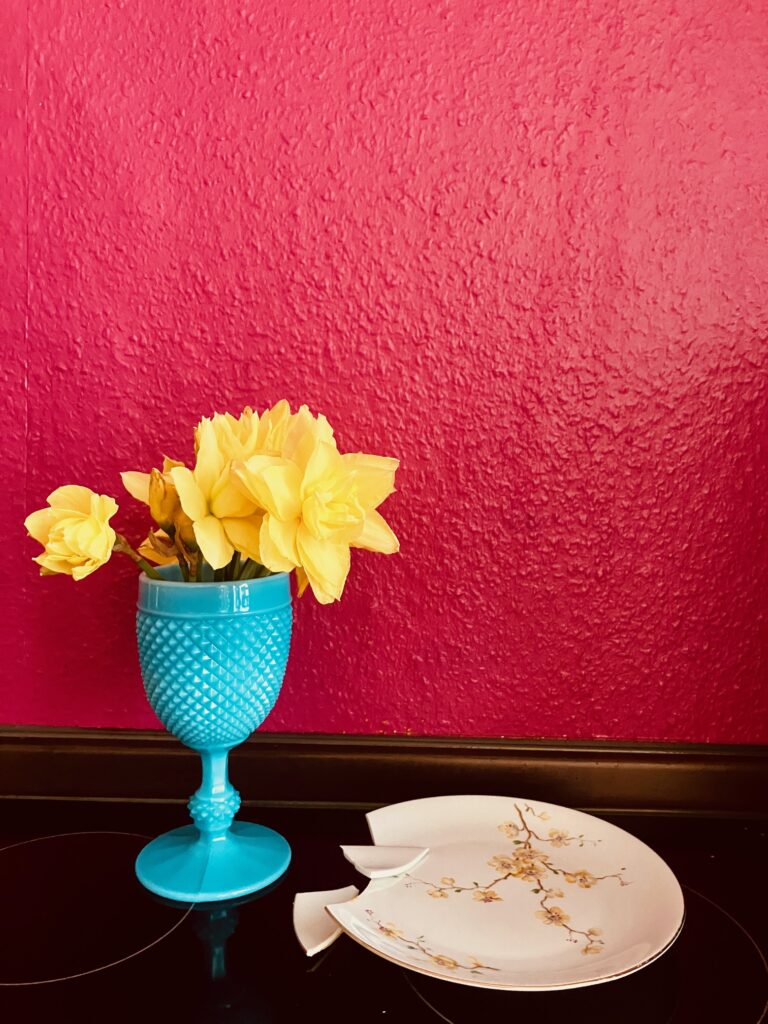 Blauer Pokal mit gelben Narzissen vor roter Wand, daneben ein weißer, gebrochener Teller mit floralem gelbem Muster