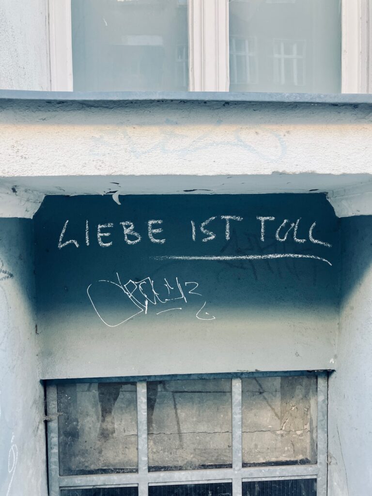 Aufschrift mit weißer Kreide mit dem Text 'LIEBE IST TOLL' in Großbuchstaben auf einer dunklen Wand über einem Kellerfenster